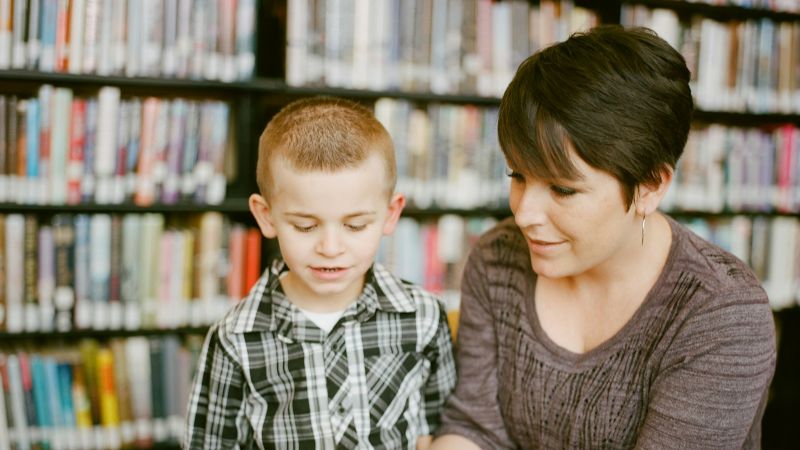 A woman and a young boy read a book together in front of library shelves