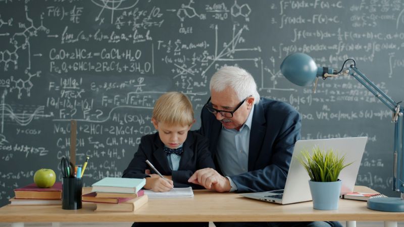 A white-haired teacher helps a young student with a math problem at a desk in front of a chalkboard