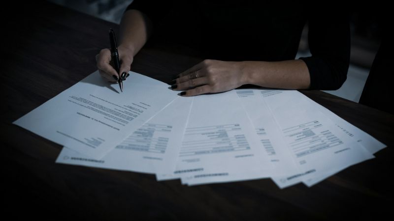 A person marks up a contract with a pen, with pages fanned out across a dark wooden table